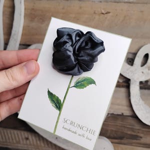 Peut inclure: Un chouchou en tissu satiné gris foncé en forme de rose, fixé sur une carte de présentation rectangulaire blanche avec une illustration aquarelle verte d'une tige de rose avec des feuilles et le texte SCRUNCHIE handmade with love.