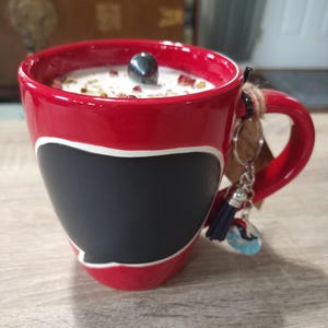 May include: Red ceramic mug with a black chalkboard design and a white rim. The mug has a small black stone on top of the white candle wax inside. A silver charm with a blue tassel hangs from the handle.