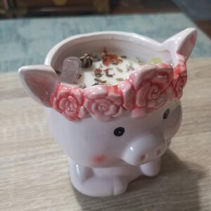 May include: A white ceramic pig candle holder with a pink floral crown and a white candle inside. The candle has dried flowers and crystals embedded in it.