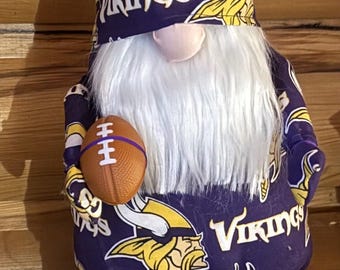 May include: Purple and yellow gnome with a white beard holding a football. The gnome is wearing a hat with the Minnesota Vikings logo and purple and white shoes.