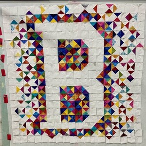 May include: A quilt made of colorful fabric squares, each square is made of smaller triangles. The quilt is arranged in a pattern of squares and triangles, with a white background. The quilt is pinned to a white wall.