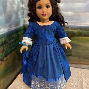 May include: A doll in a blue historical dress with white lace trim on the sleeves and skirt. The dress features a floral pattern and a matching blue purse. The doll has brown hair and blue eyes.