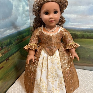 May include: A doll with brown curly hair adorned with white and gold feathers, wearing a brown and white gown with a floral pattern, ruffled sleeves, and a white beaded necklace. The gown features a fitted bodice and a full, pleated skirt.