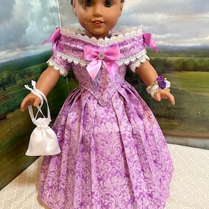 May include: A doll wearing a lavender dress with a floral pattern. The dress features a lace-trimmed neckline, a pink bow, and a matching purse. The doll also has a floral bracelet and a hairstyle with bangs.