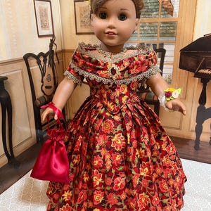 May include: A doll wearing a red floral print dress with a matching red satin purse. The dress features a lace-trimmed neckline and a jeweled brooch. The doll has a yellow and pink feather hair accessory.