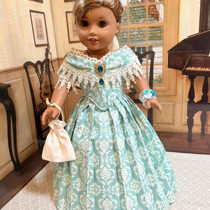 May include: A doll with brown hair styled in an updo, wearing a light blue and white damask print gown with a lace-trimmed off-the-shoulder neckline, a central blue gemstone brooch, a white wrist corsage, and holding a small white drawstring pouch.