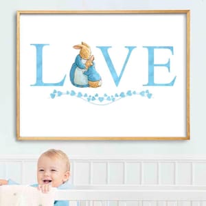 May include: A framed print with the word "LOVE" in light blue letters. The "O" is replaced by a watercolor illustration of two rabbits. A garland of blue hearts is below the word. The frame is a light wood color.