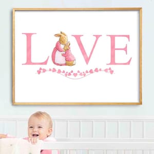 May include: A framed print with the word "LOVE" in pink capital letters. The "O" is replaced by a watercolor illustration of two bunnies. A string of pink hearts decorates the bottom. The frame is gold.