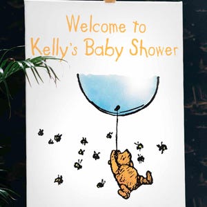 Puede incluir: Un cartel blanco con el texto "Welcome to Kelly's Baby Shower" en letras naranjas. Una ilustraci&oacute;n de dibujos animados de Winnie the Pooh sujetando un globo azul con abejas volando a su alrededor.