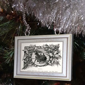 May include: A black and white vintage illustration of a horse-drawn carriage decorated with bells, framed in a silver frame with a holly border. The text below the image reads "decked with bells, which ring merrily over the snow".