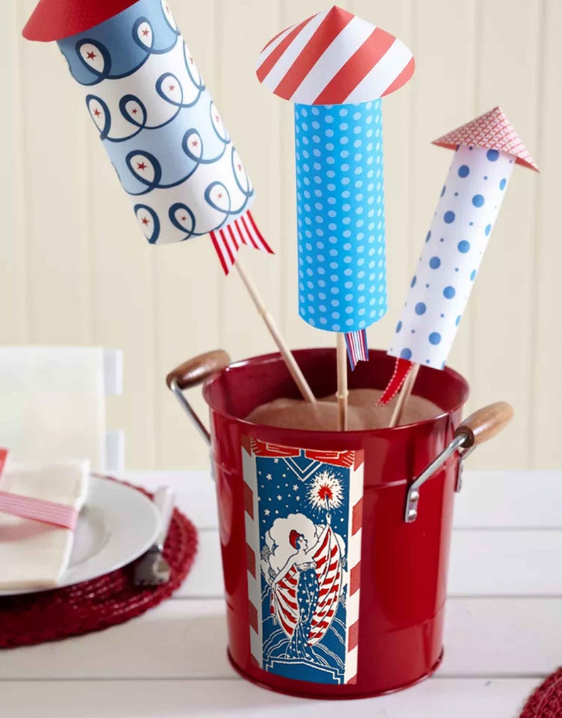 Peut inclure: Trois d&eacute;corations de p&eacute;tards en papier dans un seau en m&eacute;tal rouge. Les p&eacute;tards sont d&eacute;cor&eacute;s de motifs rouge, blanc et bleu. Le seau a une &eacute;tiquette rouge, blanche et bleue avec une femme tenant un drapeau.