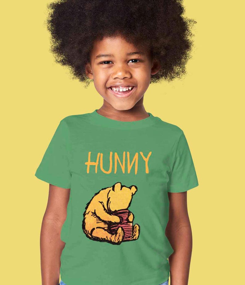Puede incluir: Una camiseta verde con una imagen de dibujos animados de Winnie de Pooh sosteniendo un tarro de miel. El texto "HUNNY" est&aacute; impreso sobre la imagen.