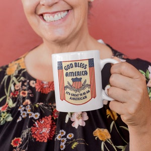 May include: A white ceramic mug with a red, white, and blue God Bless America design. The mug has a handle and the text "It's Great to Be an American".