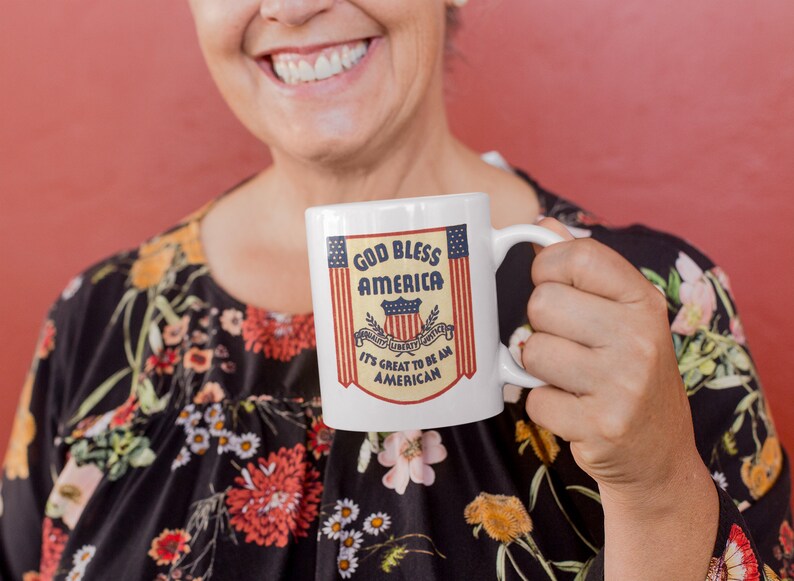May include: A white ceramic mug with a red, white, and blue God Bless America design. The mug has a handle and the text "It's Great to Be an American".