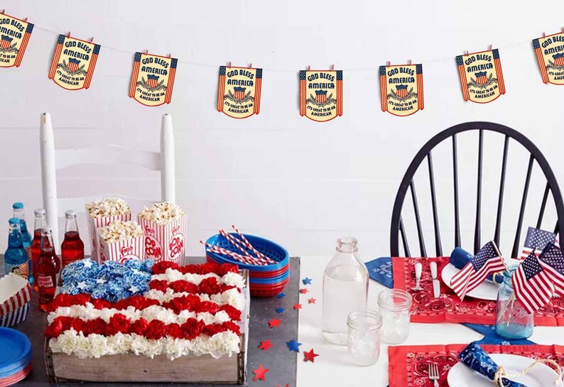 May include: A table setting for a patriotic party with red, white, and blue decorations. The table is decorated with a red, white, and blue floral arrangement, popcorn in striped containers, and red and blue bandana napkins. A string of bunting with the text "God Bless America" hangs above the table.