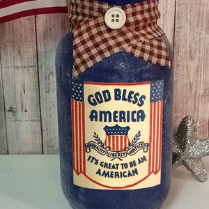 May include: A blue glass jar with a red, white, and blue fabric bow and a label that reads "God Bless America" with a shield and the words "Equality, Liberty, Justice" and "It's Great to Be an American".
