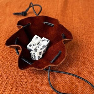 Puede incluir: Un saco de dados de cuero marrón con cierre de cordón, que contiene cuatro dados blancos con puntos negros. El saco está abierto, revelando los dados en su interior. El fondo es una tela naranja texturizada.
