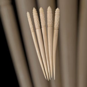 Peut inclure: Ensemble de six épingles à cheveux de couleur ivoire. Chaque épingle présente un design effilé avec un sommet décoratif sculpté. Les sommets présentent divers motifs, dont des hachures croisées et des lignes horizontales. Les épingles sont disposées en éventail.