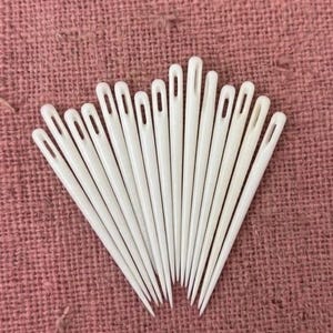 May include: A collection of white sewing needles arranged in a fan shape. Each needle has a long, slender body with a small eye at the top. The needles are set against a textured, pinkish-red fabric background, creating a contrast in color and texture.