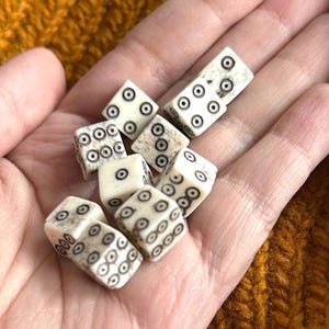 K&ouml;nnte beinhalten: Eine Sammlung elfenbeinfarbener W&uuml;rfel mit schwarzen Punktmarkierungen, die in einer Hand gehalten werden. Die W&uuml;rfel sind w&uuml;rfelf&ouml;rmig, wobei jede Seite eine unterschiedliche Anzahl von Punkten aufweist. Die W&uuml;rfel werden wahrscheinlich f&uuml;r Spiele verwendet.