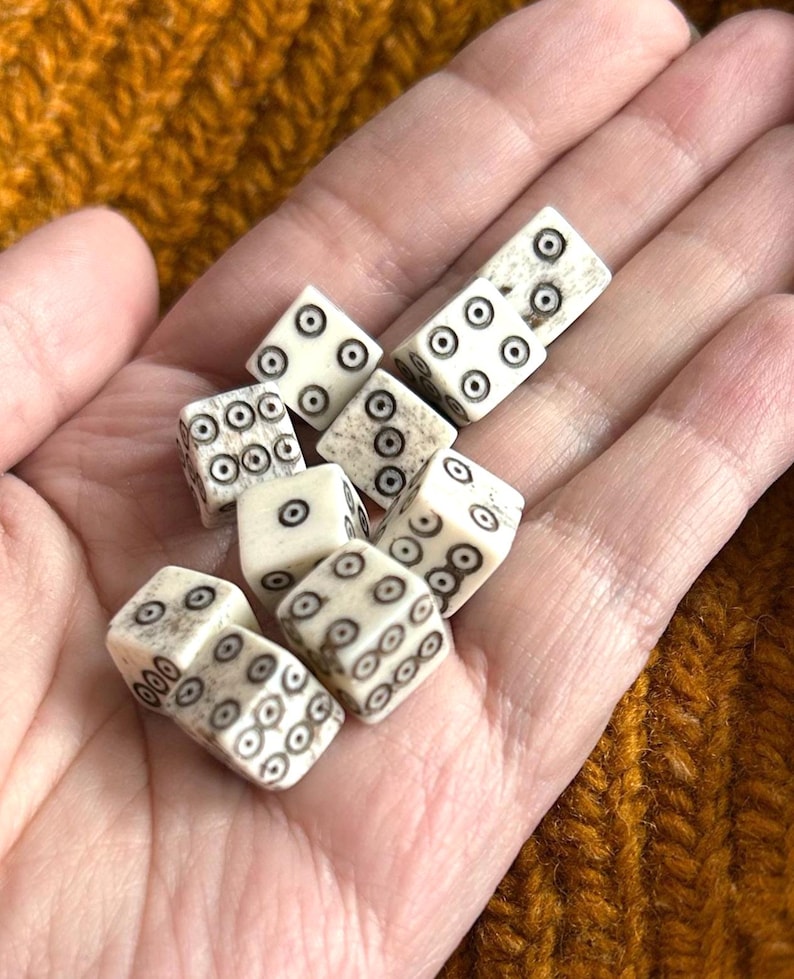 K&ouml;nnte beinhalten: Eine Sammlung elfenbeinfarbener W&uuml;rfel mit schwarzen Punktmarkierungen, die in einer Hand gehalten werden. Die W&uuml;rfel sind w&uuml;rfelf&ouml;rmig, wobei jede Seite eine unterschiedliche Anzahl von Punkten aufweist. Die W&uuml;rfel werden wahrscheinlich f&uuml;r Spiele verwendet.