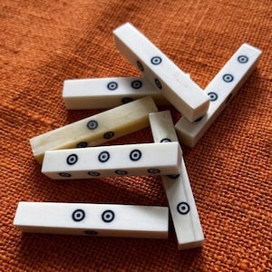 Könnte beinhalten: Elfenbeinfarbene rechteckige Spielsteine mit schwarzen und blauen kreisförmigen Markierungen. Die Teile sind auf einer orangefarbenen, strukturierten Oberfläche verstreut, was auf ein Brettspiel oder eine ähnliche Aktivität hindeutet.