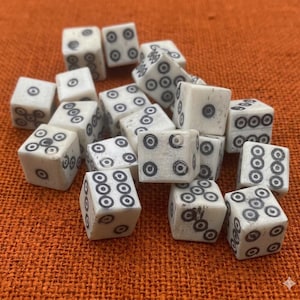 Puede incluir: Un conjunto de dados de color marfil con marcas circulares negras. Los dados están dispersos, mostrando diferentes combinaciones de números. Un accesorio de juego clásico.
