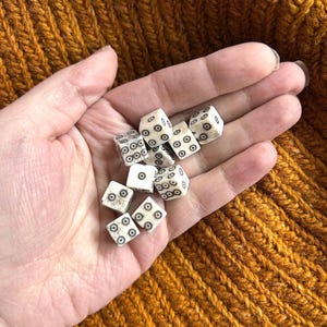 K&ouml;nnte beinhalten: Eine Handvoll kleiner, cremefarbener W&uuml;rfel mit schwarzen Punktmarkierungen. Die W&uuml;rfel sind verstreut und zeigen verschiedene Zahlen. Der Hintergrund ist ein strukturierter, senffarbener Strickstoff.