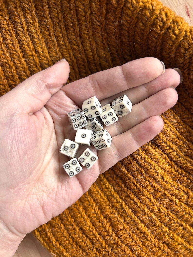 K&ouml;nnte beinhalten: Eine Handvoll kleiner, cremefarbener W&uuml;rfel mit schwarzen Punktmarkierungen. Die W&uuml;rfel sind verstreut und zeigen verschiedene Zahlen. Der Hintergrund ist ein strukturierter, senffarbener Strickstoff.