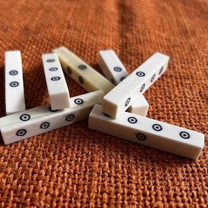 Op de afbeelding: Een verzameling rechthoekige, ivoorkleurige spelstukken met zwarte concentrische cirkelmarkeringen. De stukken liggen verspreid op een getextureerd, oranje-bruin oppervlak, wat wijst op een spel of knutselitem.