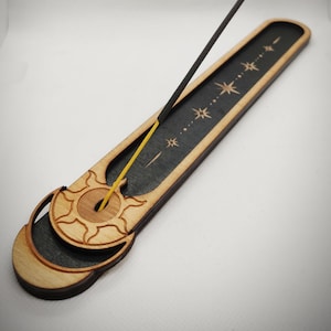 May include: A wooden incense holder with a black and wood finish. The holder features a sun and moon design with a line of stars. A black incense stick is placed in the holder.