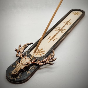 May include: Wooden incense burner with a black and white design featuring a skull with antlers and a Norse symbol. The incense burner is designed to hold a stick of incense.