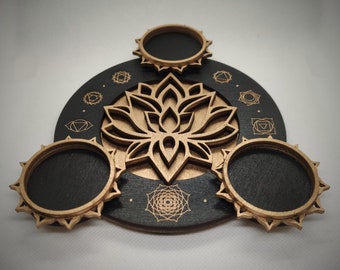 Puede incluir: Una bandeja de madera negra con un diseño de flor de loto y siete símbolos de chakras. La bandeja tiene tres hendiduras circulares para sostener objetos.