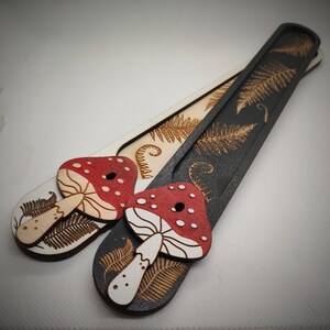 Puede incluir: Dos marcapáginas de madera con un diseño de helecho. Un marcapáginas es negro con una seta blanca y el otro es blanco con una seta roja.