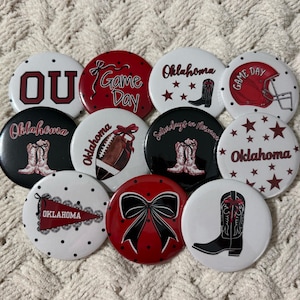 May include: A collection of round buttons with Oklahoma-themed designs. The buttons feature text like "OU", "Game Day", and "Oklahoma", along with images of a football, cowboy boots, and a football helmet. The color scheme is primarily red, white, and black.