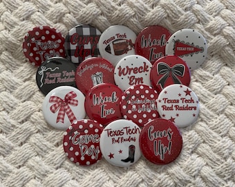 May include: A collection of round buttons with various designs and text, primarily in red, white, and black. The buttons feature phrases like "Guns Up!" and "Wreck 'Em," along with the words "Texas Tech" and football-related graphics.