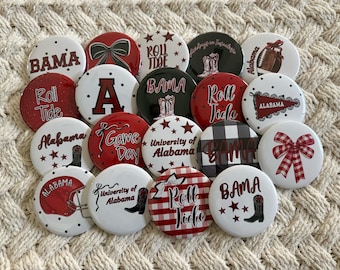May include: A collection of round buttons with various designs and text, primarily in red, white, and black. The buttons feature phrases like "BAMA," "Roll Tide," and "Game Day," along with images of a football, boots, and bows. The buttons are arranged on a textured, off-white surface.