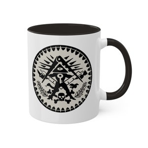 May include: White ceramic mug with a black interior and handle. The mug features a black and grey circular design with an eye, key, and other symbols. The design is surrounded by a decorative border.