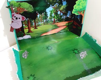 May include: A green cardboard box diorama depicting a forest scene with a koala hanging from a tree branch. The box is open and the inside is decorated with a green grassy field and a dirt path leading into the forest. The text "FOREST DIORAMA" is printed on the front of the box.