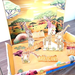 May include: A giraffe diorama with a savanna scene. The diorama features several giraffe figures, trees, and grass. The background depicts a sunset with a yellow and orange sky. The text "GIRAFFE DIORAMA DIJITAL FILE" is visible.