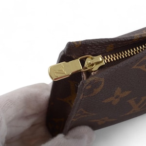 May include: Brown and tan patterned pouch with a gold zipper. The zipper pull has a gold logo. The pouch is held by a gloved hand. The pattern is a repeating design of four-petal flowers.