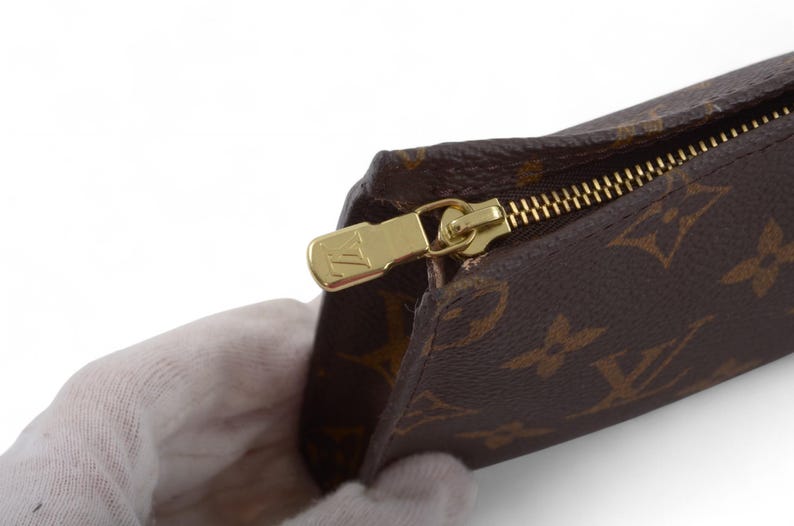 May include: Brown and tan patterned pouch with a gold zipper. The zipper pull has a gold logo. The pouch is held by a gloved hand. The pattern is a repeating design of four-petal flowers.