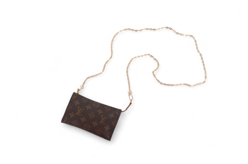 May include: A brown pouch-style bag with a gold chain strap. The bag features a repeating pattern of brown logos on a brown background. The gold chain is long and thin, suitable for wearing the bag as a crossbody.