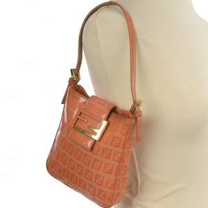 May include: A peach-colored shoulder bag with a glossy finish. The bag features a repeating pattern of the letter "F" and a gold-colored buckle. The strap is a matching peach color. The bag is displayed on a mannequin.