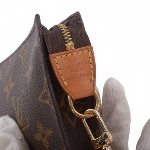 May include: Close-up of a brown and tan monogrammed clutch with a gold zipper and hardware. A leather tag reads "LOUIS VUITTON PARIS made in France". The clutch is held by a gloved hand.