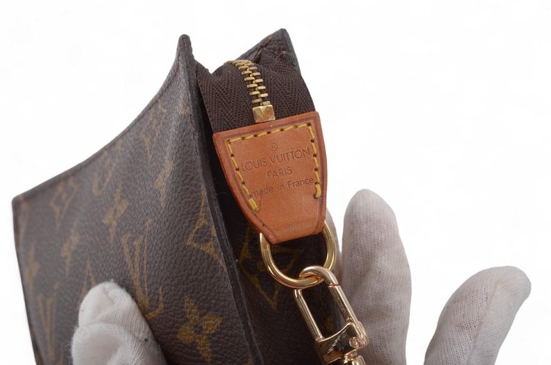 May include: Close-up of a brown and tan monogrammed clutch with a gold zipper and hardware. A leather tag reads "LOUIS VUITTON PARIS made in France". The clutch is held by a gloved hand.