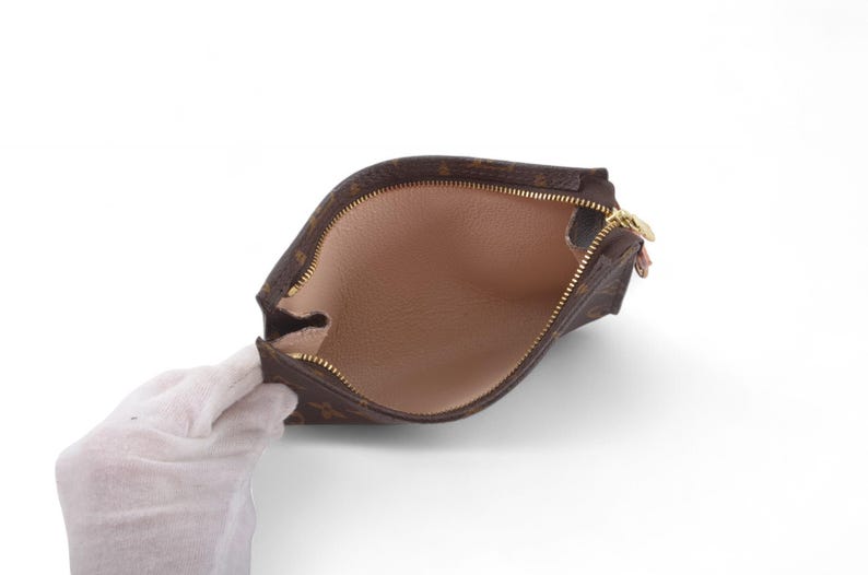May include: Brown and tan zippered pouch with a gold-colored zipper. The interior is a light tan color. The pouch is held open by a gloved hand.