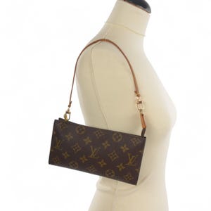 Könnte beinhalten: Braune, rechteckige Handtasche mit einem sich wiederholenden Muster aus ineinandergreifenden Buchstaben und floralen Formen. Die Tasche hat einen dünnen, hellbraunen Lederriemen und goldfarbene Beschläge. Die Tasche wird auf einer Schaufensterpuppe präsentiert.