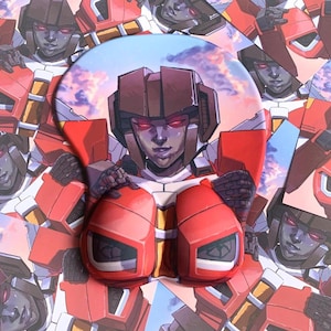 Puede incluir: Alfombrilla de ratón roja y blanca con reposamuñecas, con temática de Transformers. El diseño presenta una imagen central de un personaje robot con un casco rojo y marrón, rodeado por un patrón repetido del mismo personaje.