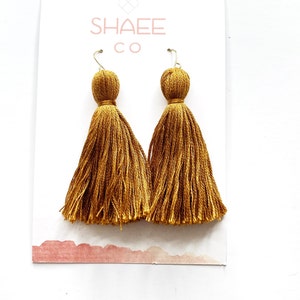 May include: A pair of gold tassel earrings with a long, thin tassel design. The earrings are on a white card with the text "Shaee Co."
